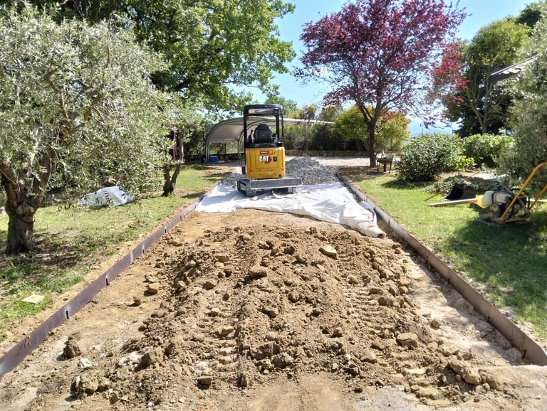 Yellow excavator spreading soil in a garden bed surrounded by olive trees and flowering shrubs