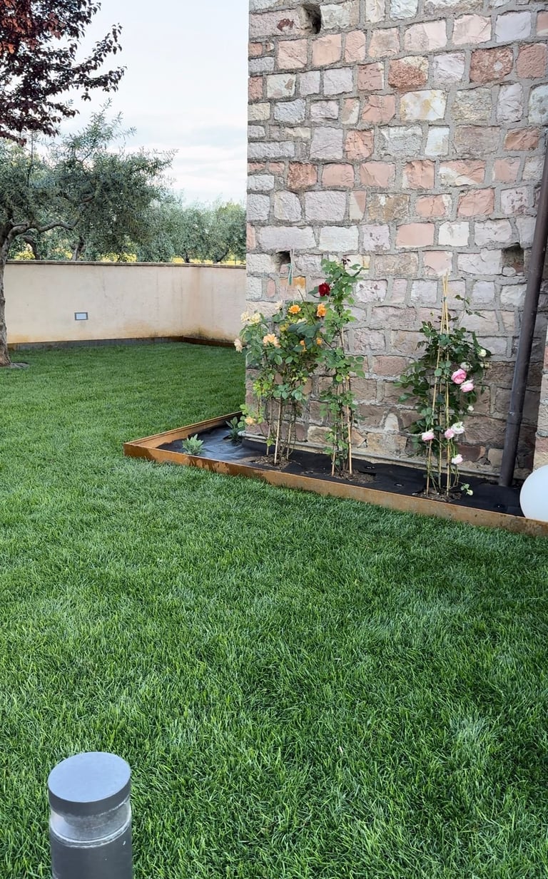 Giardino con prato verde rigoglioso, aiula rialzata con piante colorate contro muro di mattoni