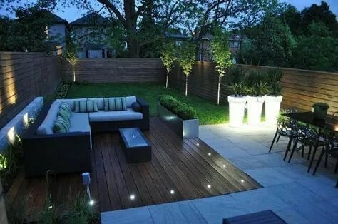 Giardino moderno con deck in legno illuminato, divano blu esterno e mobili scuri al crepuscolo