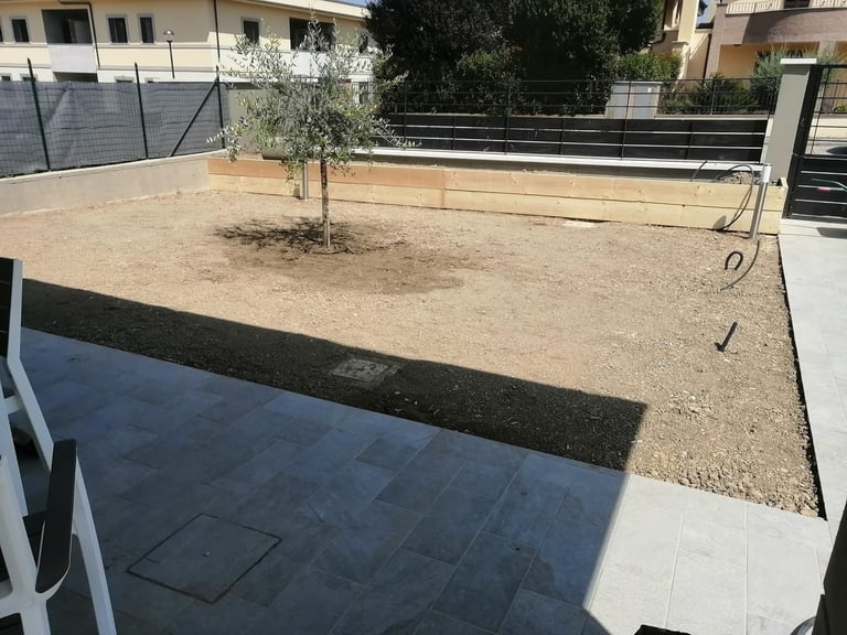 Construction site showing a patio area with dark paving stones next to a sandy yard with a young tree and metal fencing