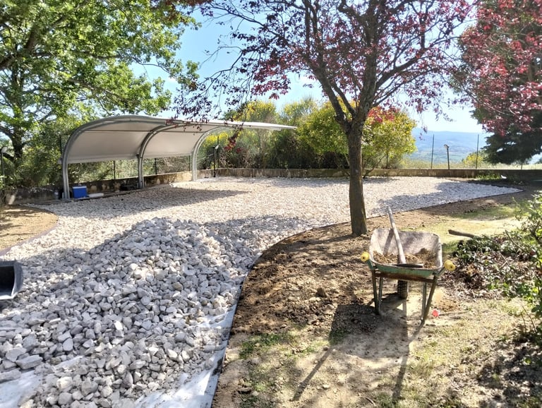 Outdoor recreational area with covered pavilion, gravel pathways, trees, and picnic area under sunny blue sky