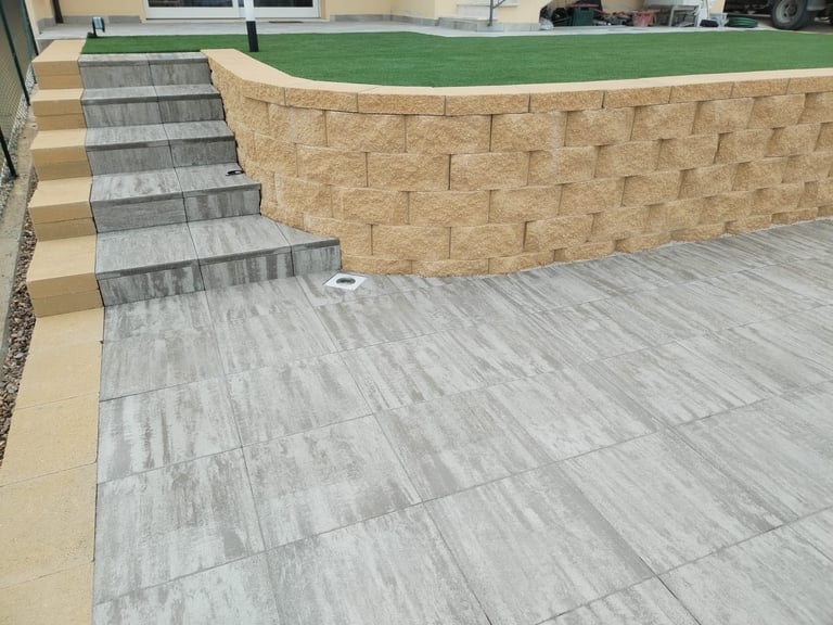 Backyard patio with gray tile flooring, stone retaining wall, stairs, and artificial green lawn area with house in background