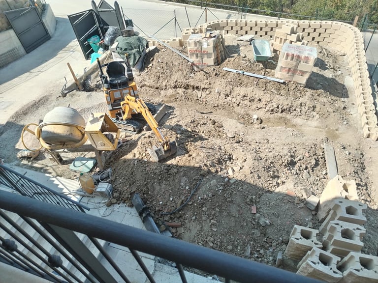 Overhead view of a construction site with yellow excavator, cement mixer, and piles of materials on sandy ground