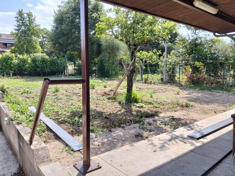 View of a backyard garden with young trees, grass, and a metal railing in the foreground under a wooden awning structure